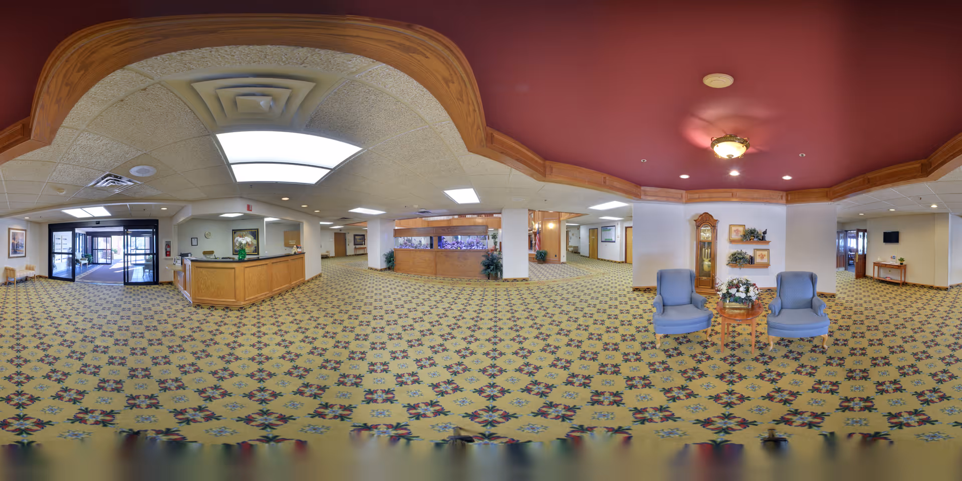 Wide panoramic view of the interior lobby area of Heritage Oaks facility featuring patterned carpet flooring, a wooden reception desk, a large fish tank, two blue armchairs with a small round table and floral arrangement between them, a grandfather clock, framed artwork on the walls, and multiple doorways leading to other parts of the building.