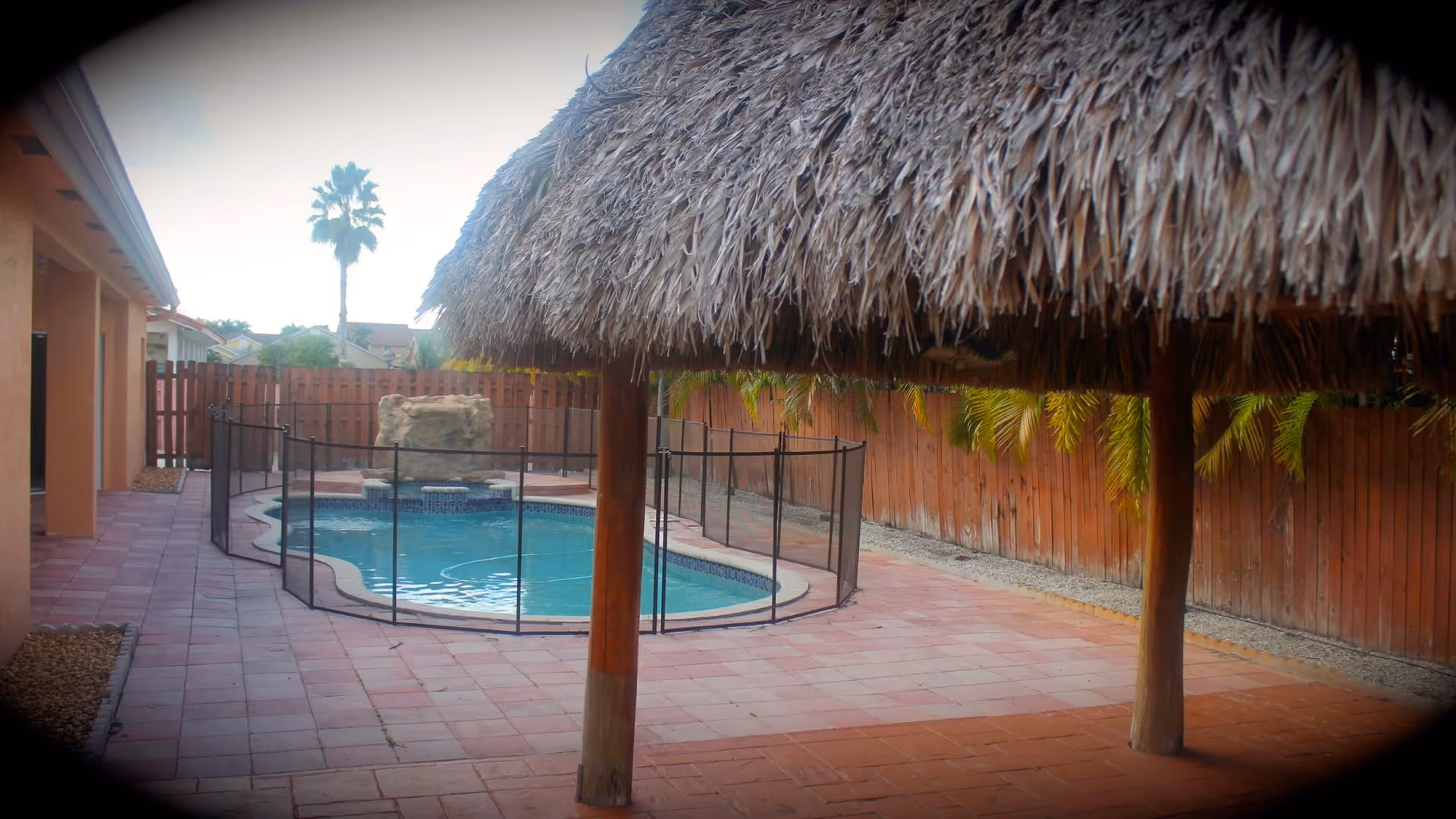 Outdoor area with a small swimming pool surrounded by a safety fence, a thatched roof structure with wooden posts providing shade, a red brick patio, a wooden fence, and palm trees in the background.