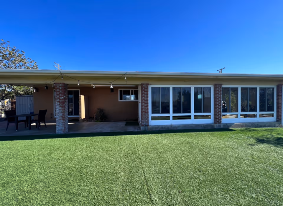 Exterior view of a single-story building with large windows and brick columns, a covered patio area with outdoor furniture, and a well-maintained green lawn under a clear blue sky.