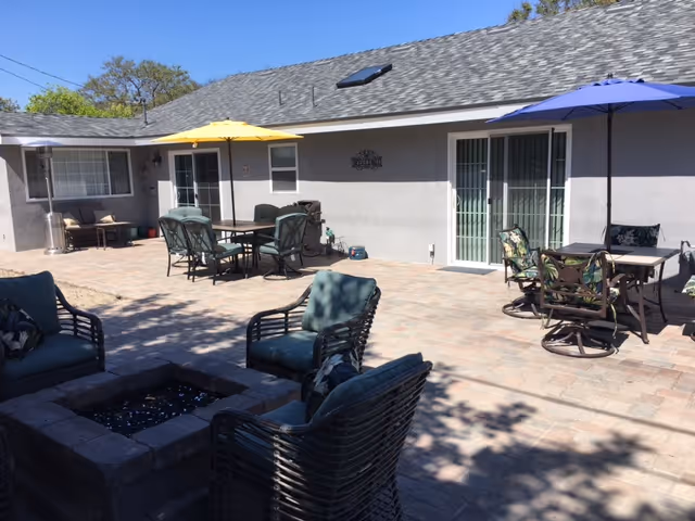 Outdoor patio area at Addie's Cottage Senior Living with multiple seating arrangements including cushioned chairs around a fire pit and tables with umbrellas. The patio is paved with bricks and attached to a single-story building with sliding glass doors and windows.