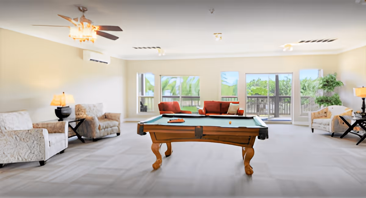 Bright communal lounge with a pool table centered, upholstered seating and lamps, and large windows/doors opening to a balcony.