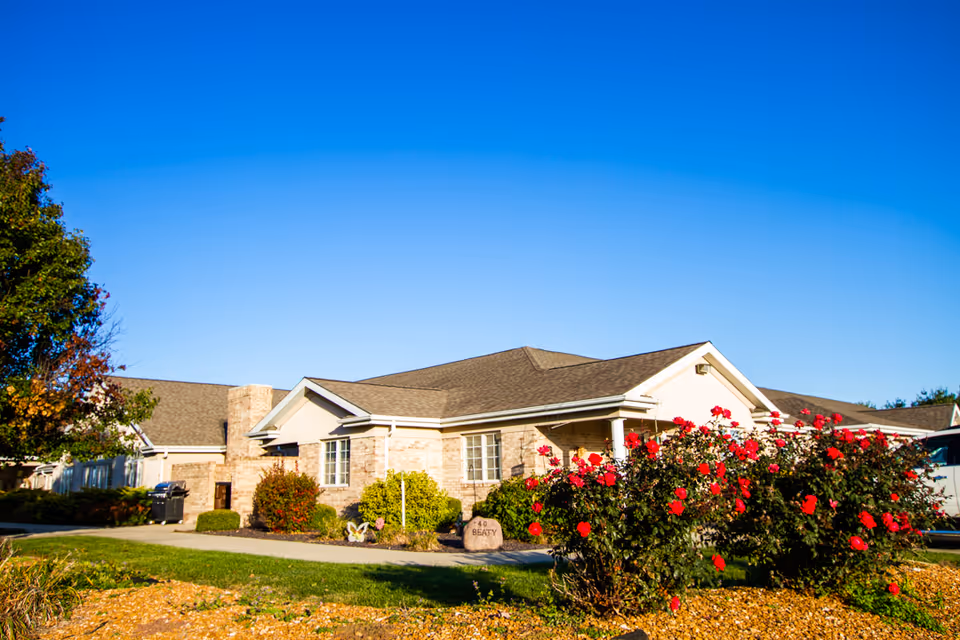 Exterior view of a single-story senior living facility building with beige brick walls and a brown roof under a clear blue sky. There are bushes with red flowers and a well-maintained lawn in front of the building.