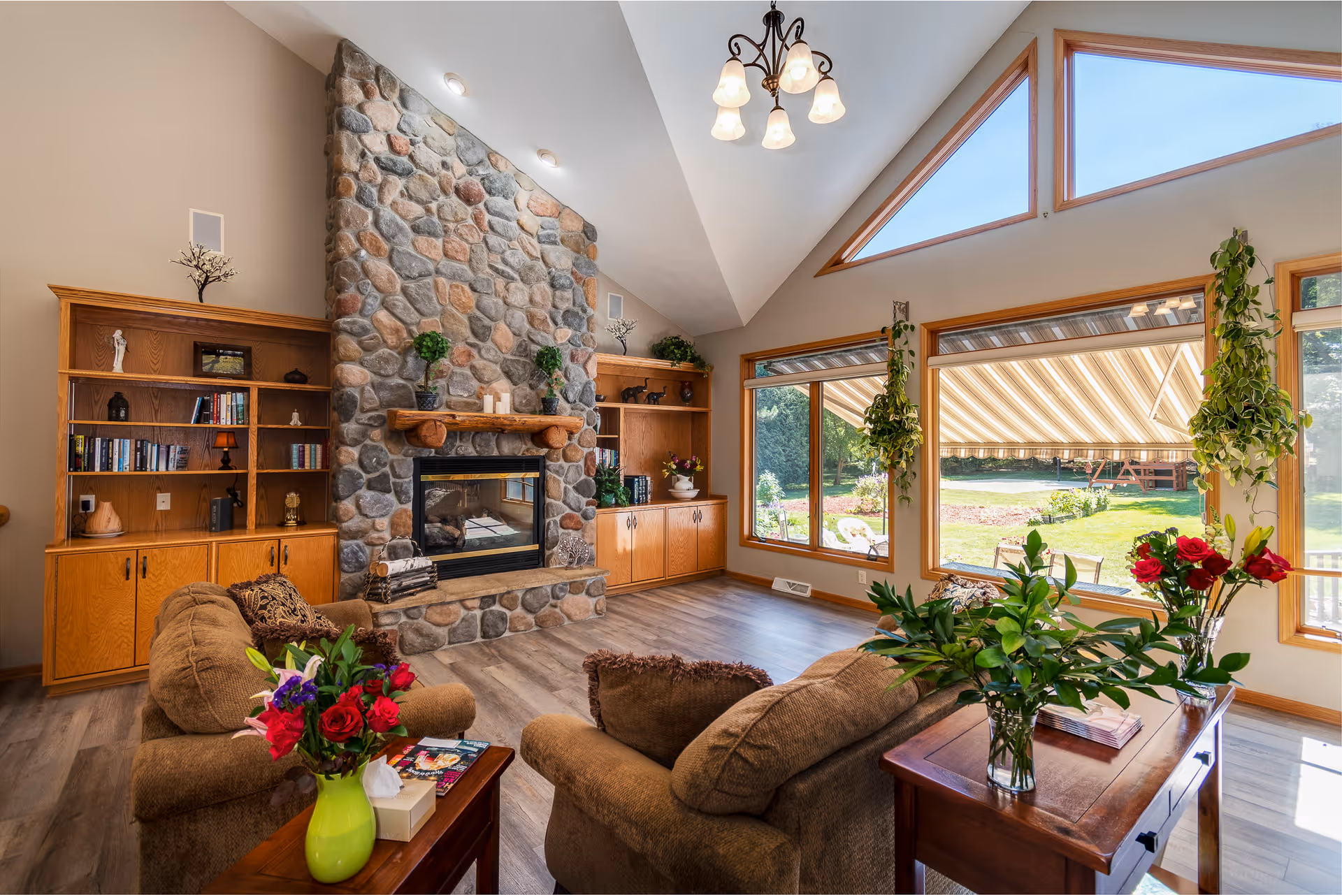 A cozy living room with a large stone fireplace flanked by wooden built-in bookshelves. Two brown armchairs face the fireplace, with a wooden side table between them holding a green vase with red and purple flowers. Large windows with wooden frames let in natural light and offer a view of a green outdoor space with a striped awning. The room has a high ceiling with a chandelier and wooden flooring.