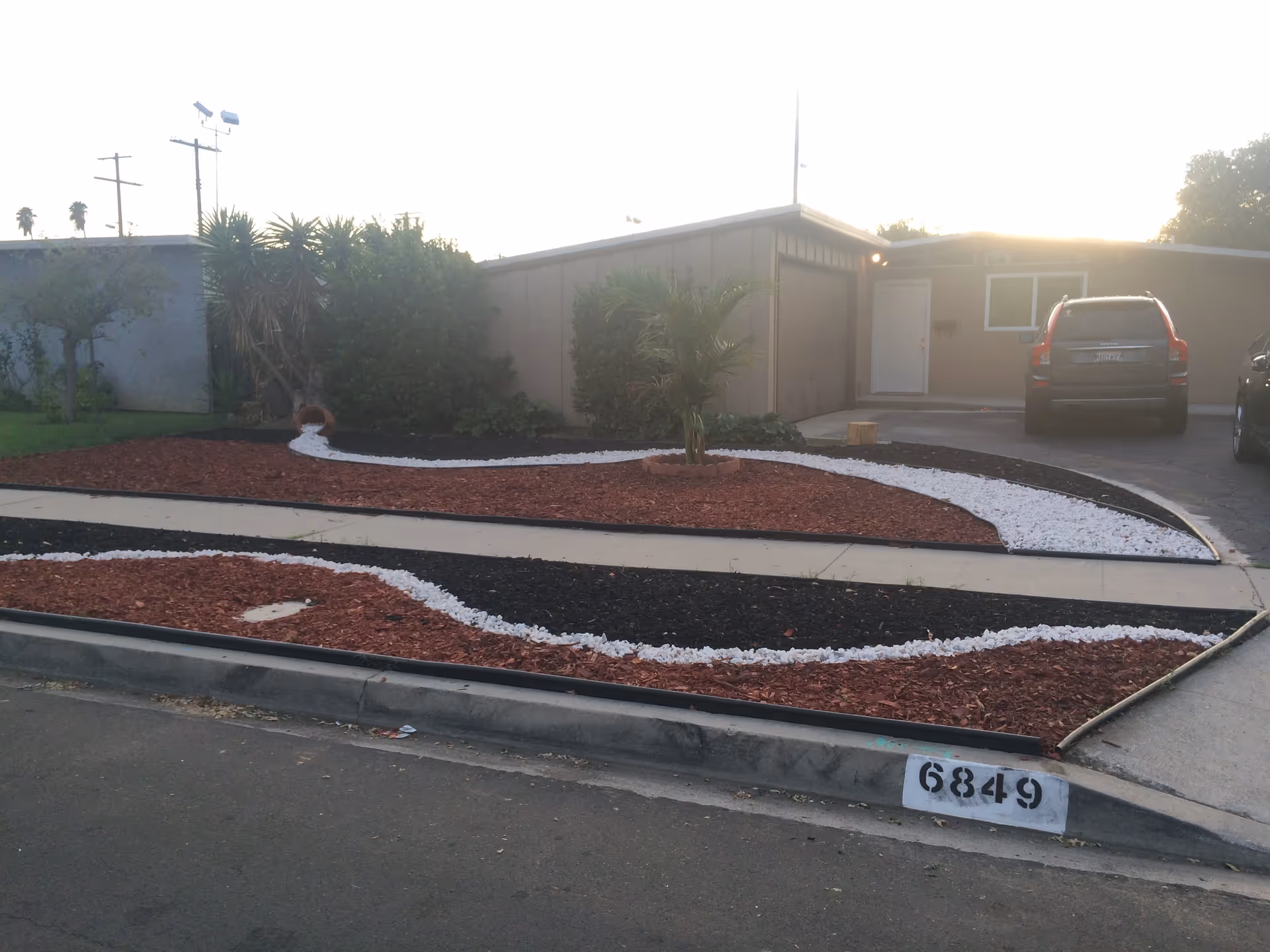 Front yard of a single-story house with a driveway and two parked cars. The yard features a decorative landscaping design with red and black mulch separated by white rocks, a small palm tree, and other plants near the house. The house has a garage and a white front door.
