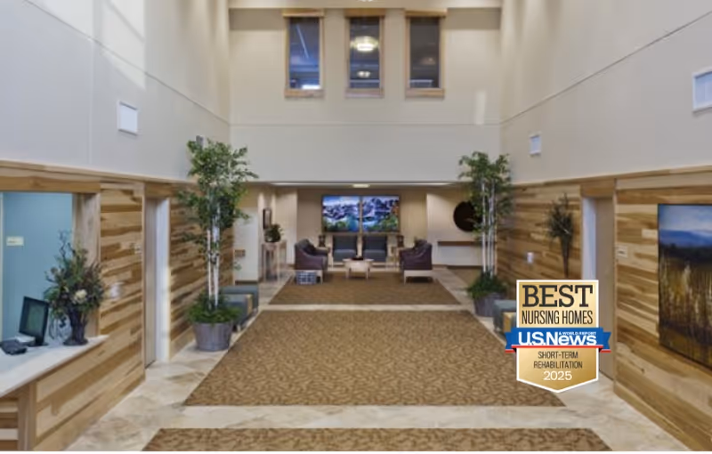 Interior view of a senior living facility lobby or common area with high ceilings, beige walls, wood panel accents, potted plants, seating area with chairs and a couch, and decorative artwork on the walls.