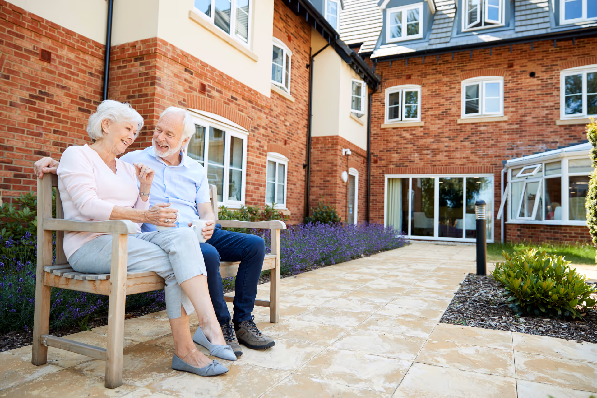 An elderly couple sitting on a wooden bench outside a brick assisted living facility, smiling and enjoying a conversation. The area is landscaped with purple flowers and green shrubs along a paved walkway.