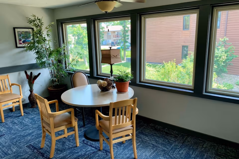 A small seating area inside Garnett Place with a round table surrounded by four wooden chairs. The room has large windows showing greenery and a neighboring building outside. There is a potted plant on the table, a lamp, a tall plant in the corner, and a framed picture on the wall.