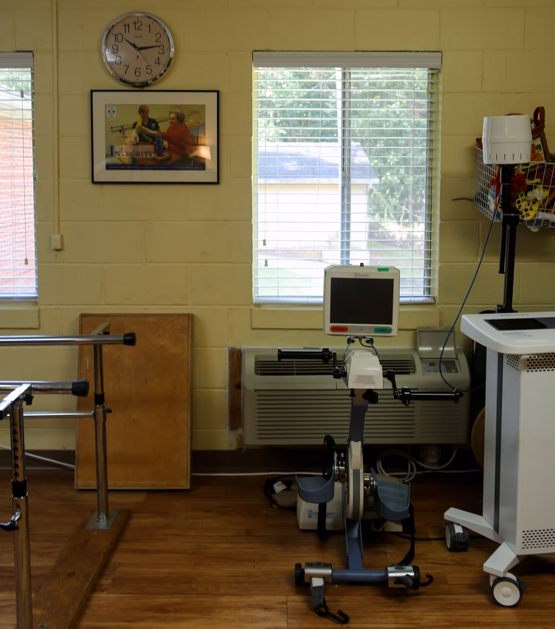 Physical therapy room with exercise equipment including a stationary pedal exerciser and parallel bars. Two windows with blinds let in natural light. A clock and a framed poster are mounted on a beige brick wall.