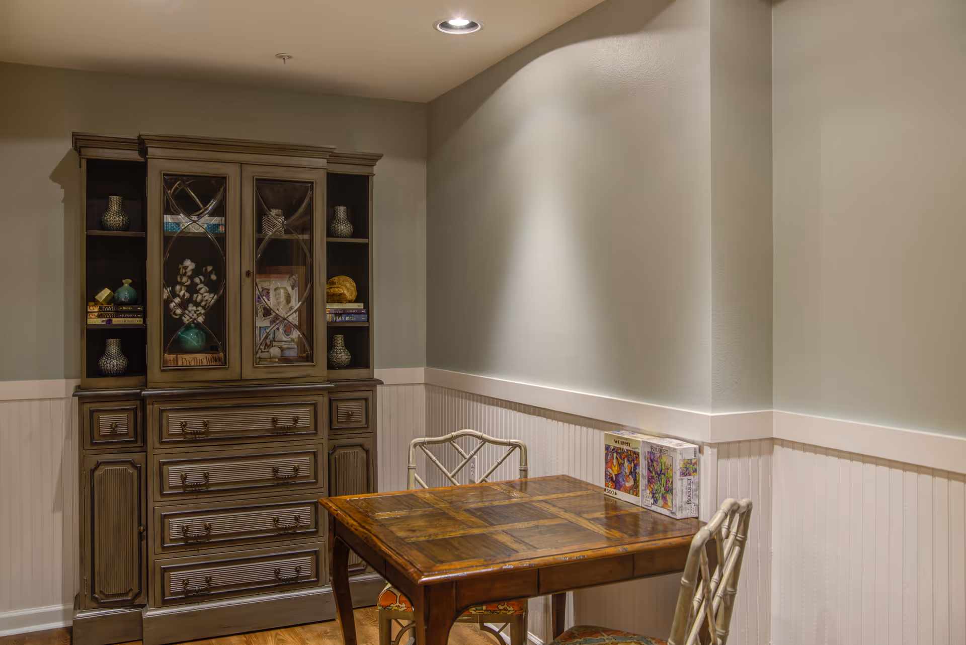 A small room with a wooden table and two chairs. Against the wall is a large wooden cabinet with glass doors displaying decorative items and books. Two puzzle boxes are placed on the table near the wall. The walls are painted light gray with white wainscoting, and the floor is wooden.
