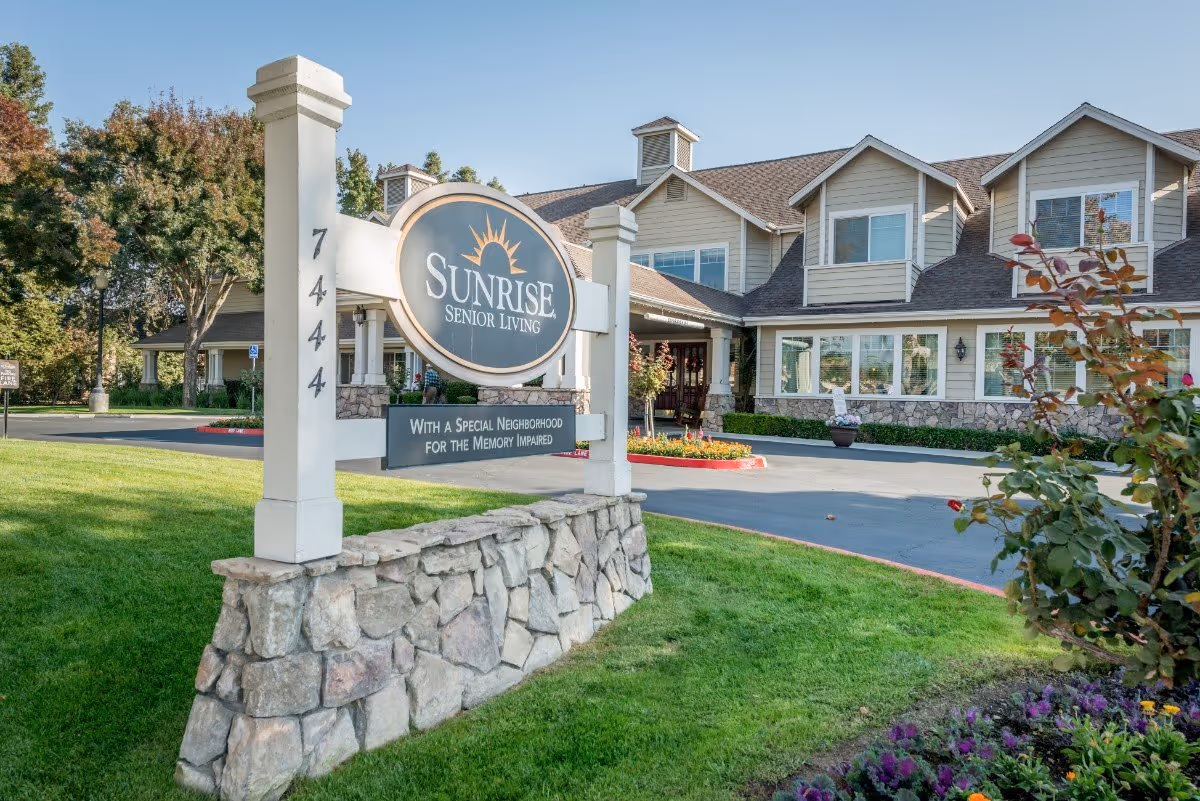 Exterior front entrance of Sunrise Senior Living with a large wooden sign, driveway, and landscaped lawn.