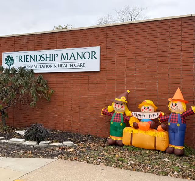 Exterior brick wall of Friendship Manor Rehabilitation & Health Care facility with a sign mounted on the wall. In front of the wall are three inflatable scarecrow decorations dressed in fall attire, holding a banner that says 'Happy Fall'. There are some plants and fallen leaves on the ground nearby.