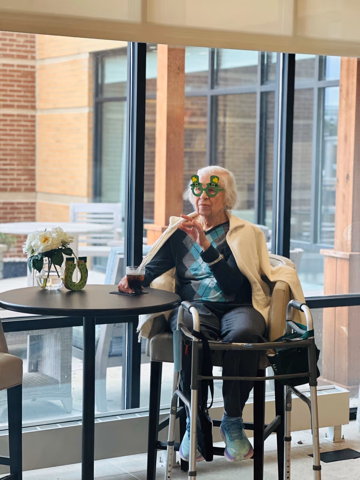 An elderly woman wearing festive green shamrock glasses sits in a chair next to a round table with a drink and a small flower arrangement. She has a walker in front of her and is indoors near large windows showing an outdoor patio area.