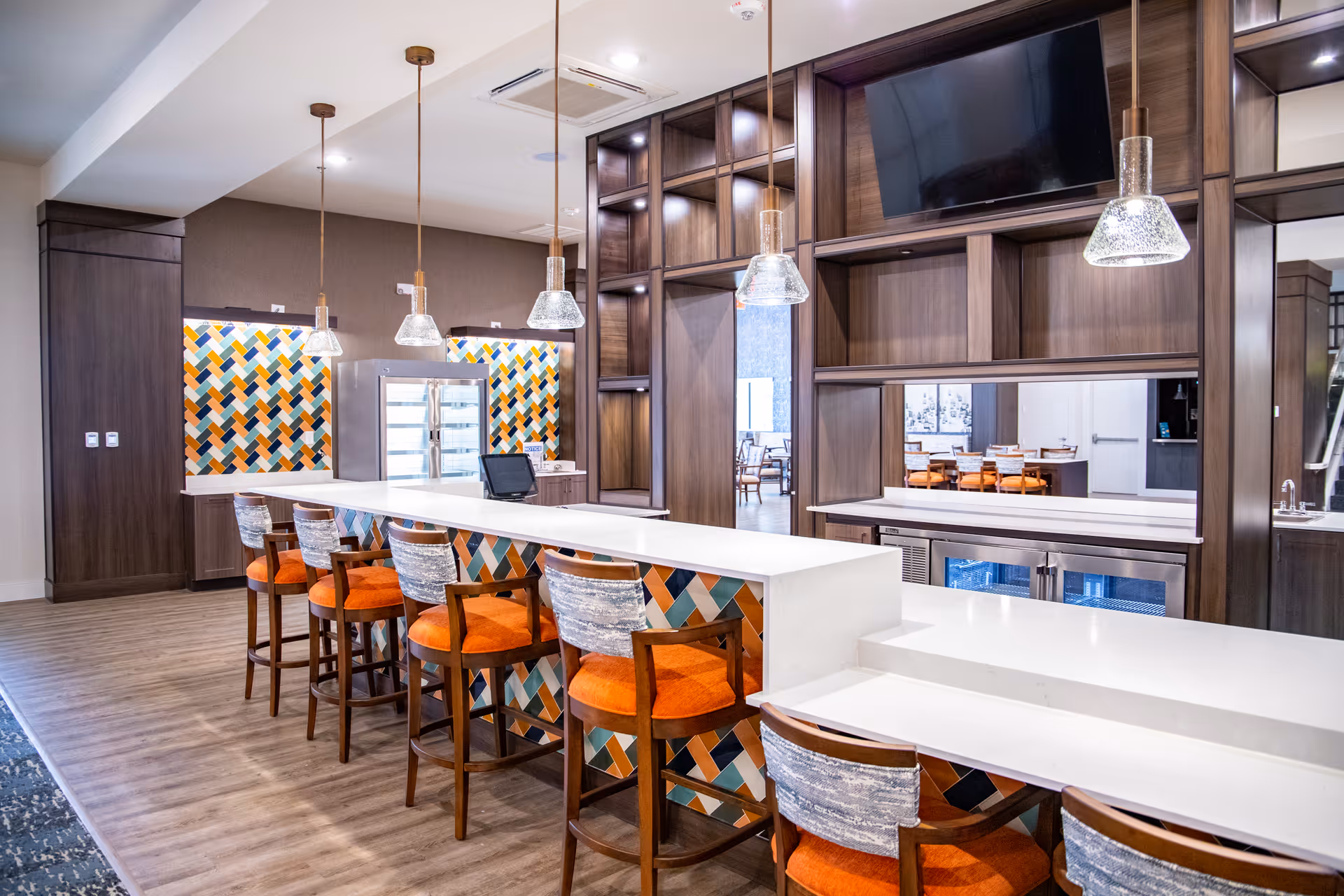 Modern dining area with a long white counter and six wooden chairs with orange cushions and patterned backs. The counter features a colorful geometric tile design on the front. Above the counter hang four pendant lights with glass shades. Behind the counter is a wall with dark wood shelving, a large flat-screen TV, and a pass-through window to another dining area with tables and chairs. The floor is wood, and the walls have a neutral tone with colorful geometric accent panels.