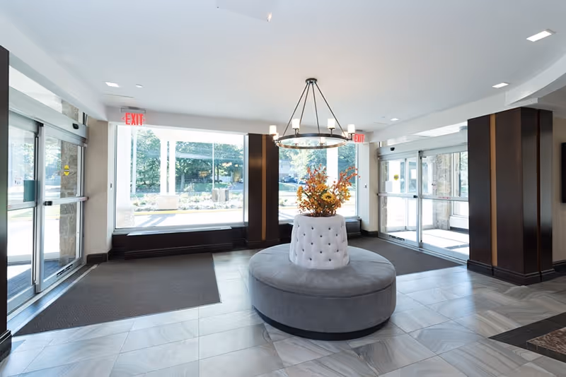 Bright and spacious entrance lobby with large glass doors and windows allowing natural light to fill the space. A round cushioned bench with a floral arrangement in the center sits under a modern chandelier. Dark wood accents frame the windows and doors, and gray tiled flooring covers the area.