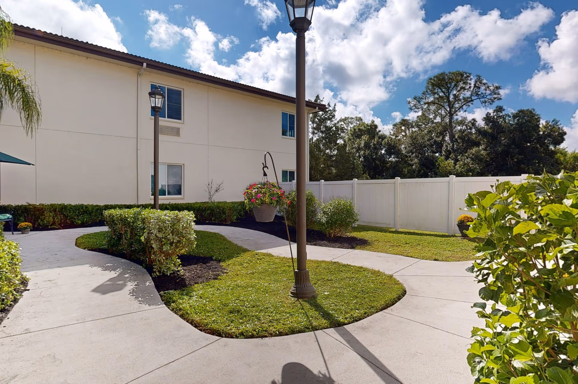 Outdoor courtyard area at The Windsor of Venice featuring a paved walkway curving around landscaped grass and bushes, a hanging flower pot on a lamp post, a white privacy fence, and a two-story building under a partly cloudy sky.