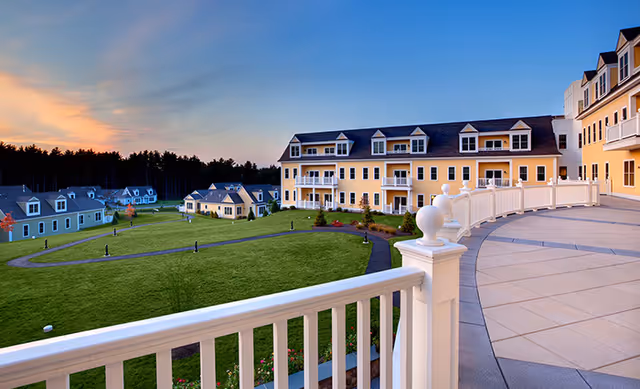 View from a curved balcony railing overlooking a landscaped lawn and yellow multi-story senior living buildings at sunset.