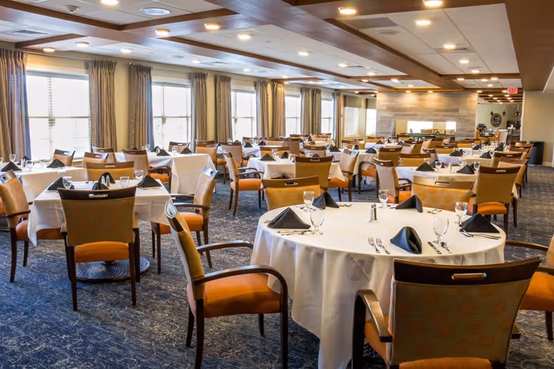 A spacious dining room with multiple tables covered in white tablecloths, each set with black folded napkins, glassware, and silverware. The room features large windows with beige curtains allowing natural light to fill the space, and wooden chairs with orange cushions surround the tables. The ceiling has recessed lighting and wooden panel accents.