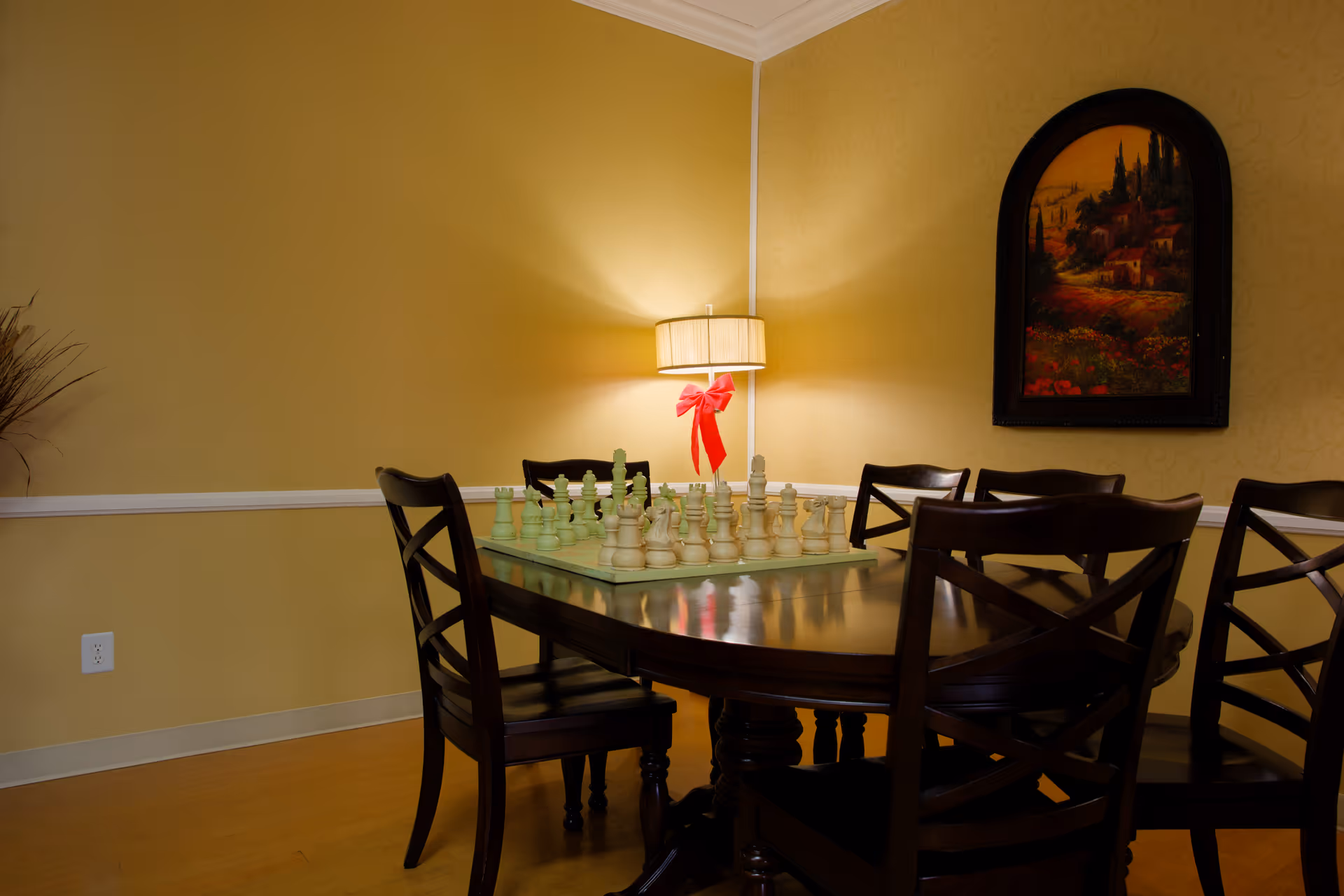 A cozy room with a dark wooden table and six matching chairs arranged around it. On the table is a large chess set. A wall lamp with a beige shade and a red bow is mounted on the wall, casting warm light. A framed painting depicting a scenic landscape hangs on the wall to the right.