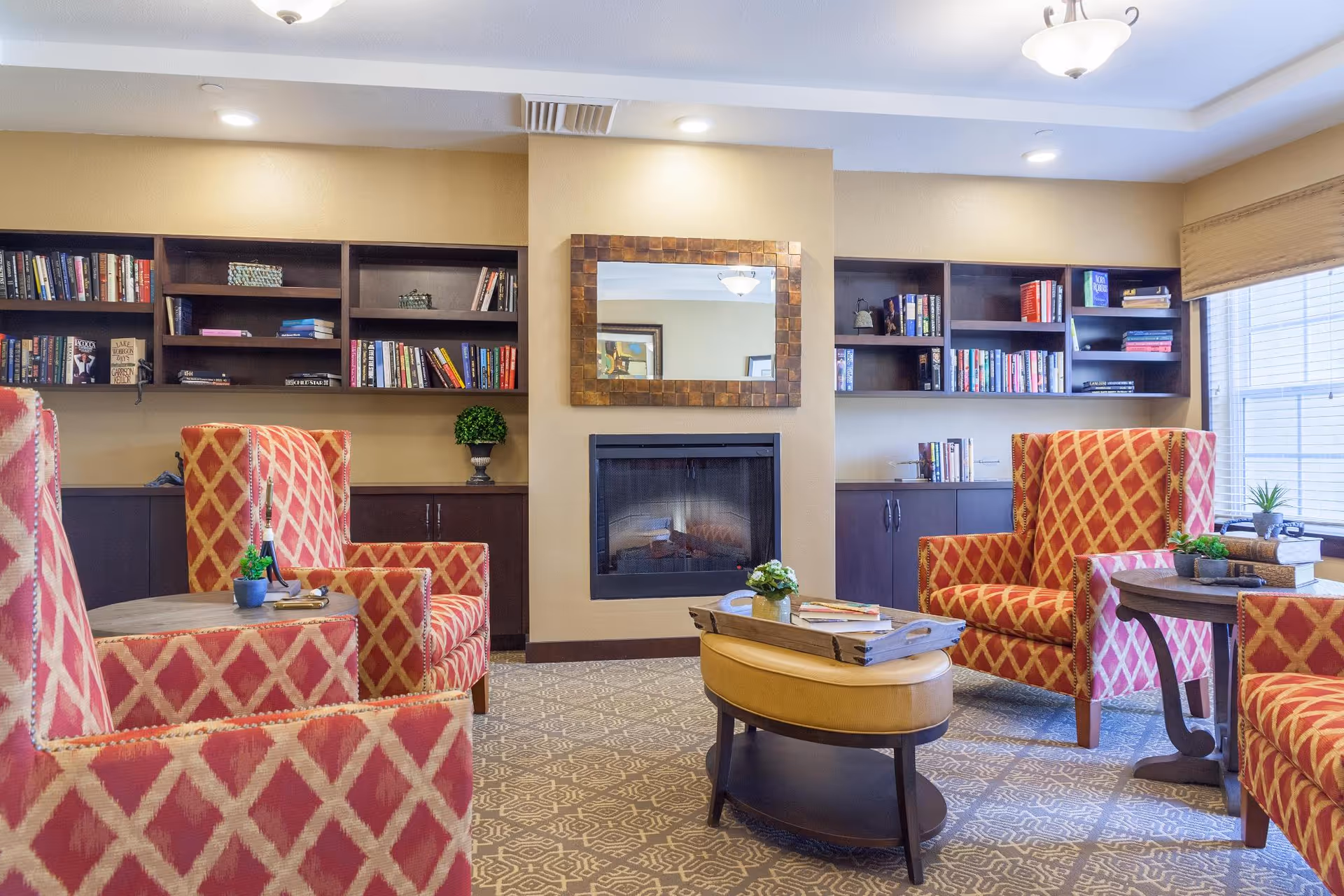 A cozy living room area with four red and gold patterned armchairs arranged around a small oval coffee table. The room features a fireplace with a decorative mirror above it, built-in dark wood bookshelves filled with books, and a large window with blinds letting in natural light.