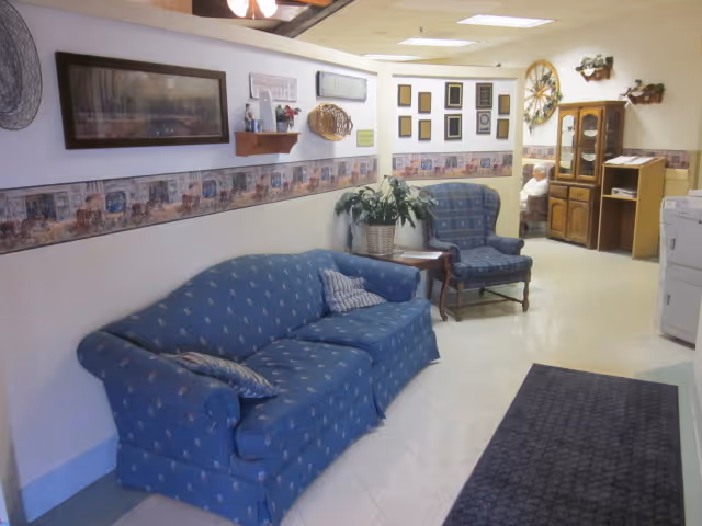 A cozy interior room with a blue patterned sofa and matching armchair along a wall decorated with framed pictures and a wallpaper border. There is a small wooden side table with a plant on it between the sofa and chair. In the background, there is a wooden cabinet and a person sitting in a chair. The floor is tiled and there is a dark rug on the floor near the entrance.