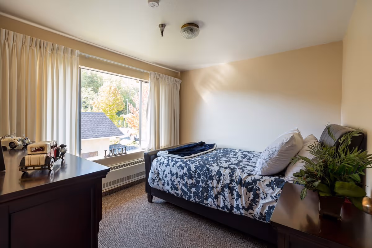 A cozy bedroom with a single bed covered in a blue and white floral bedspread, two pillows, and a folded blanket. There is a wooden dresser with model cars on it and a wooden nightstand with a green potted plant. A large window with white curtains lets in natural light and shows trees outside. The word 'LAUGH' is displayed on the windowsill.