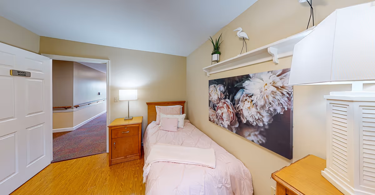 Small model bedroom with a single bed, nightstands and lamps, floral wall art, and an open door to a hallway.