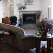A cozy living room area featuring a stone fireplace with a white mantel, decorated with plants and a metal wall art piece above it. In front of the fireplace is a floral-patterned sofa and a wooden side table with a potted plant. Additional seating and plants are visible around the room, along with a floor lamp providing soft lighting.