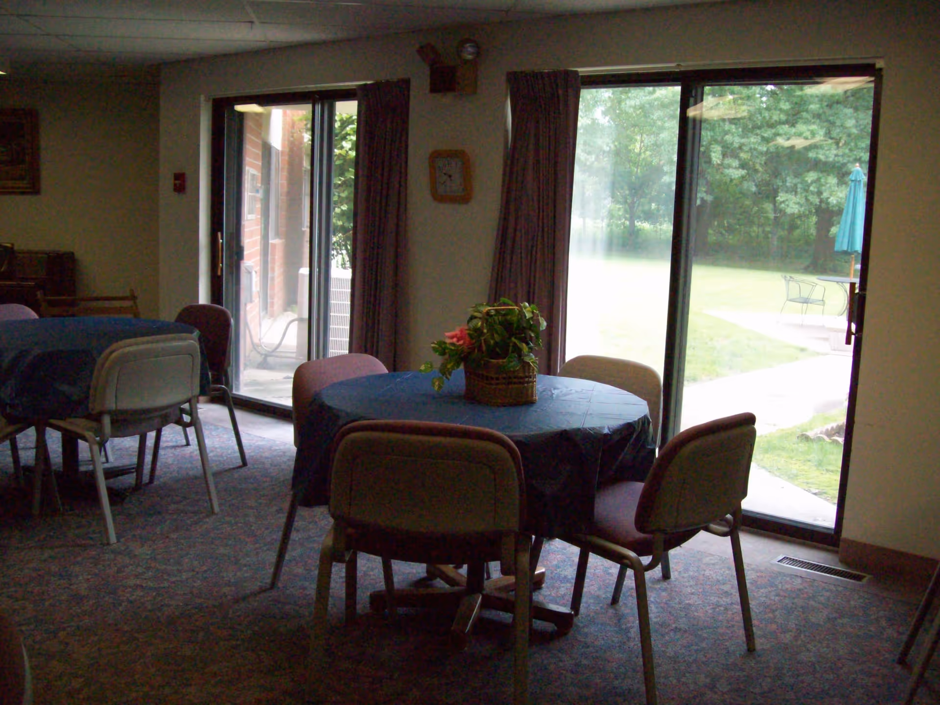 Dining/common room with round tables covered in blue cloths, chairs, and sliding glass doors opening to a grassy outdoor area.