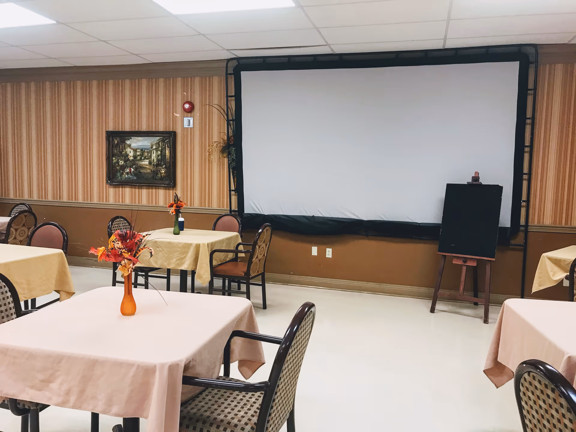 A room with several tables covered with beige tablecloths, each table having a small vase with artificial flowers. There are chairs around the tables, a large white projection screen on the wall, a framed painting, and a small blackboard on an easel. The walls have striped wallpaper and the floor is light-colored.