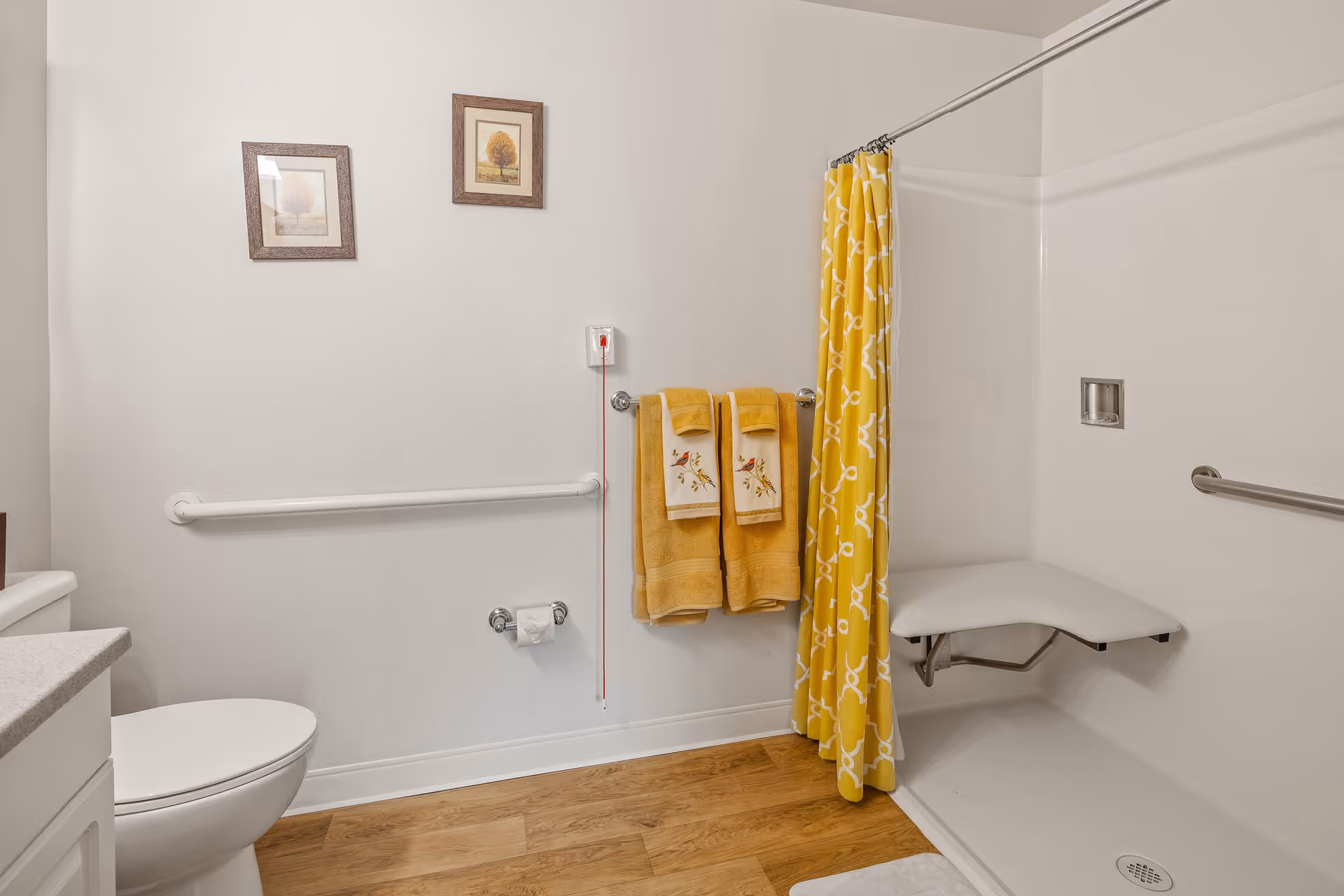 A bathroom with a white toilet, a grab bar on the wall, two yellow towels hanging on a towel rack, a yellow shower curtain with a white pattern, and a fold-down shower seat inside the shower area. The floor has wood-like flooring, and there are two framed pictures on the wall.