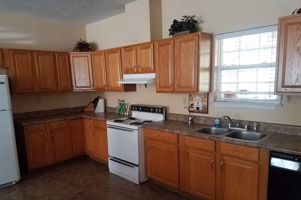 A kitchen with wooden cabinets, a white refrigerator, a white stove with an overhead vent, a double sink under a window with blinds, and a dishwasher. The countertops are brown, and there are some decorative plants on top of the cabinets.