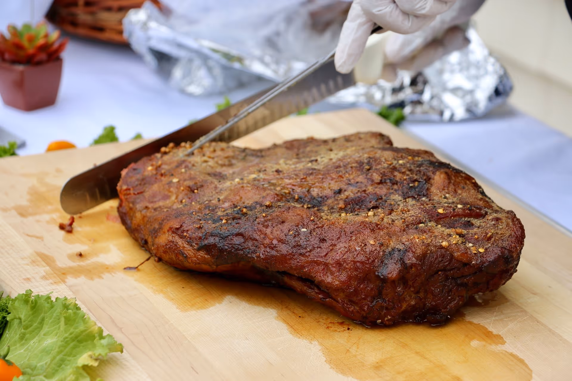 A large piece of cooked meat being sliced on a wooden cutting board with a gloved hand holding the knife. The background shows a table with a white tablecloth, some foil-covered dishes, and a small potted plant.