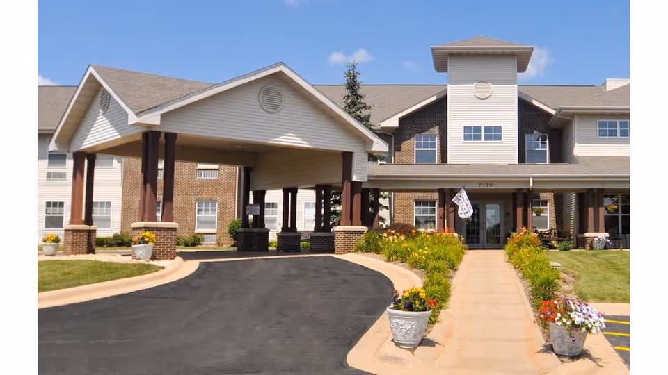 Front entrance of a senior living building with a covered drop-off, walkway, and flower planters.