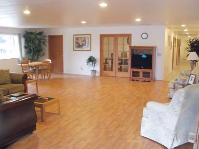 A spacious living room area with wooden flooring, featuring a brown leather sofa, a light-colored armchair, a small wooden coffee table, and a TV on a wooden stand. There is a small dining table with chairs near a window, potted plants, framed artwork on the walls, and a hallway leading to other rooms.