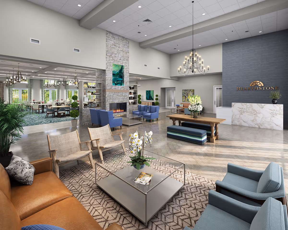 Spacious and well-lit senior living facility common area with a modern fireplace, comfortable seating including blue armchairs, wooden chairs, and a brown leather sofa. A large chandelier hangs from the ceiling, and there is a wooden table with floral arrangements. The reception desk with HearthStone logo is visible on the right, and a dining area with tables and chairs is seen through an open doorway on the left.