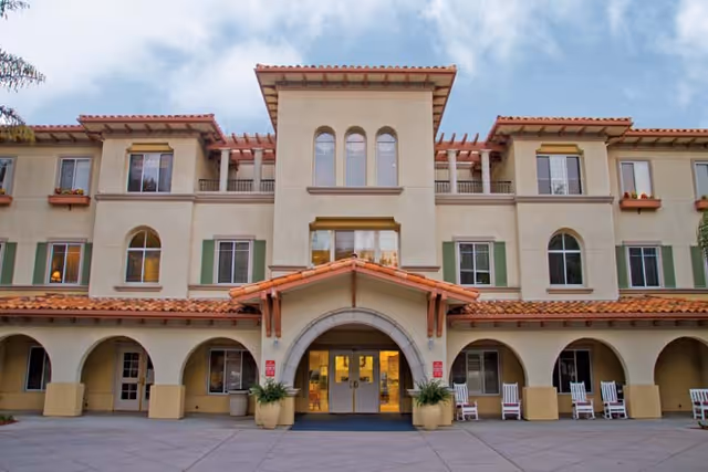 Front exterior view of a three-story senior living facility building with a tiled roof, arched entryway, multiple windows with green shutters, and several white rocking chairs on the porch area.