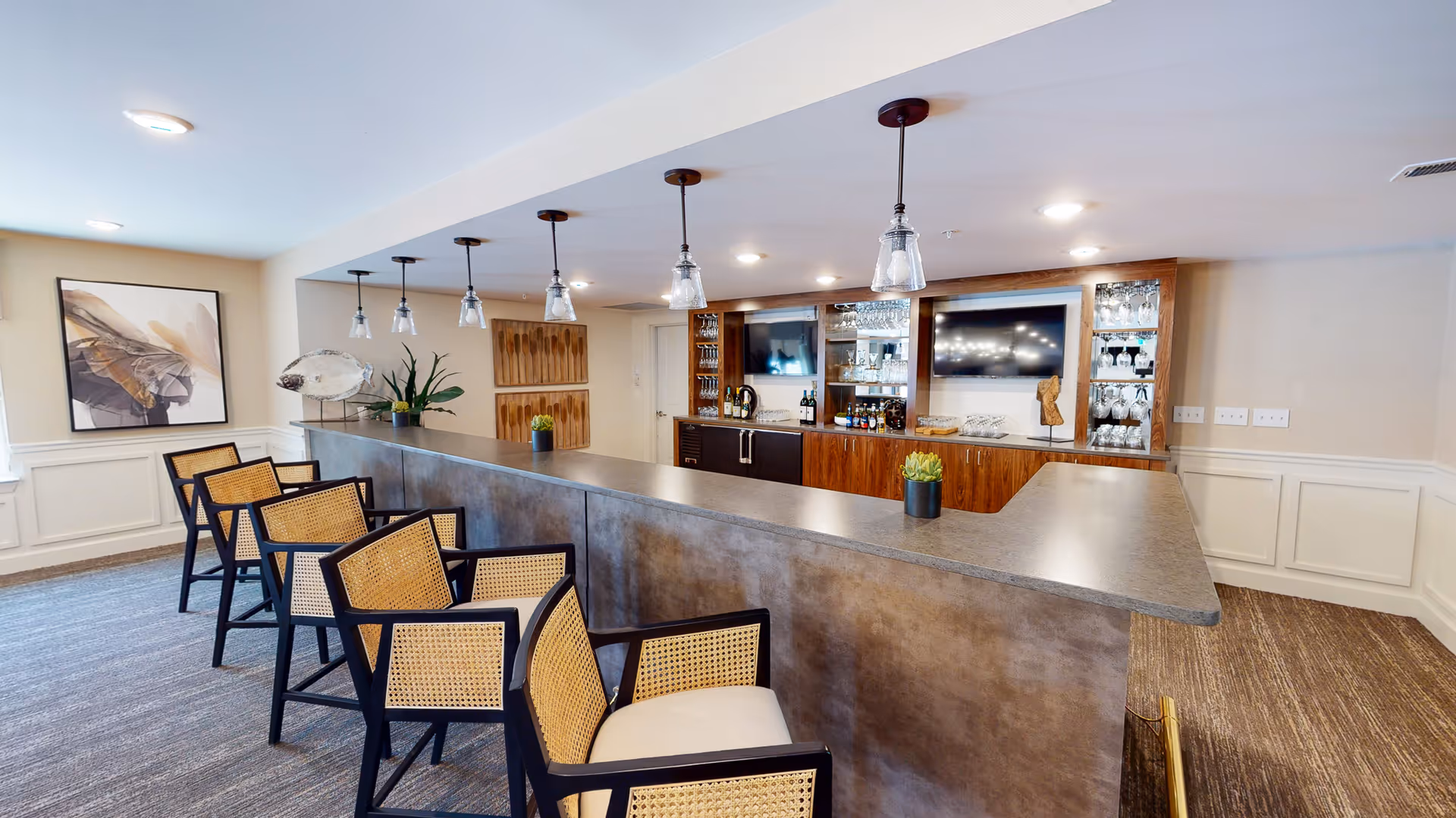 Interior view of a modern bar area in a senior living facility with a long counter, six high chairs with woven backs and cushioned seats, pendant lights hanging from the ceiling, and a wooden cabinet with glassware and two televisions mounted on the wall.