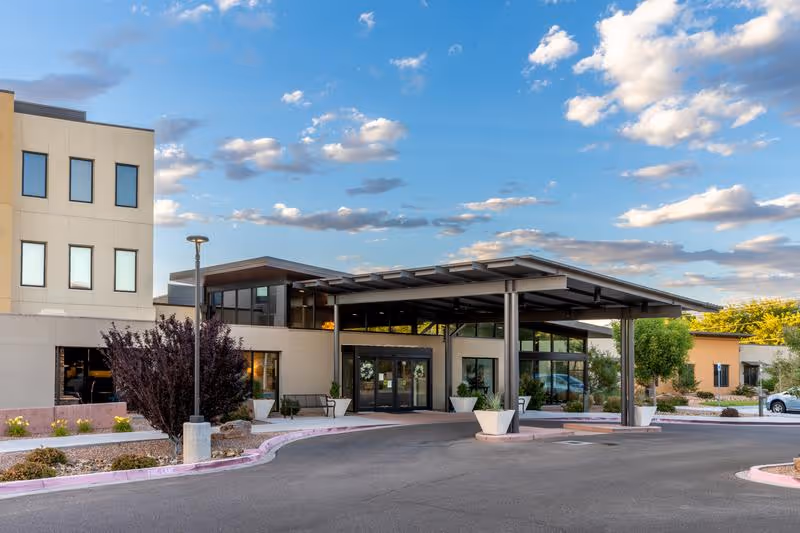 Front entrance of a modern beige building with a covered porte-cochère, driveway, and planters under a partly cloudy sky.