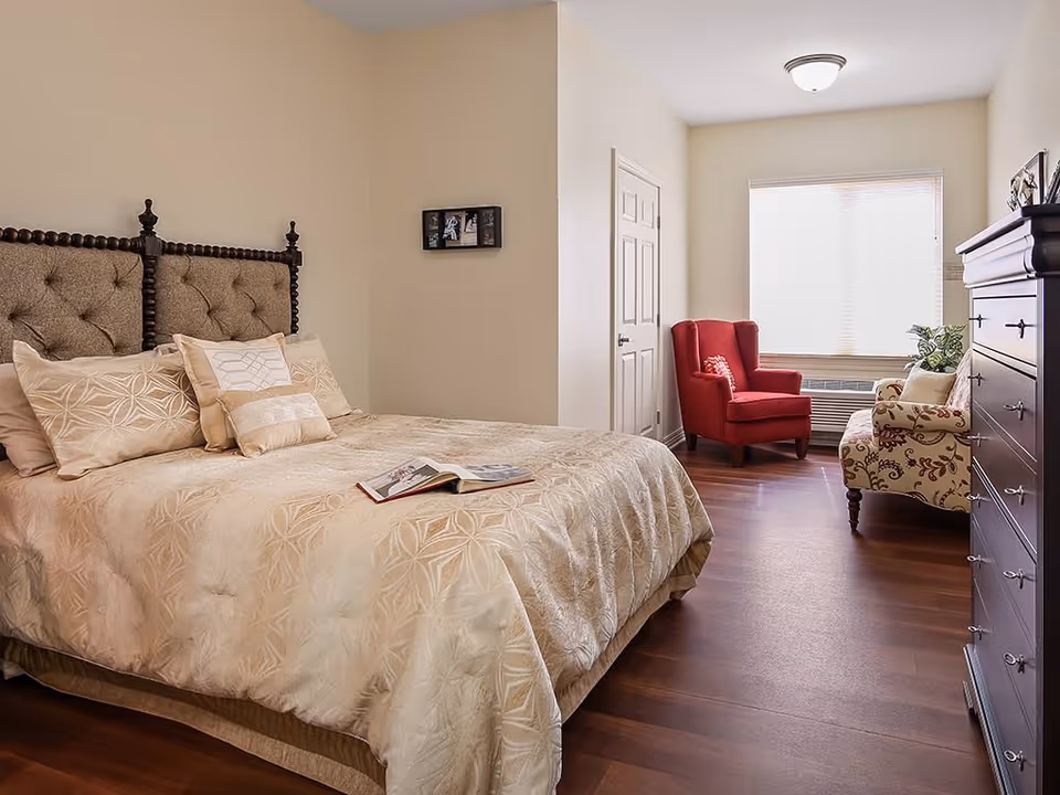 Bright furnished bedroom with a large bed and open book on it, a red armchair and patterned chair by a window, and a tall dresser.