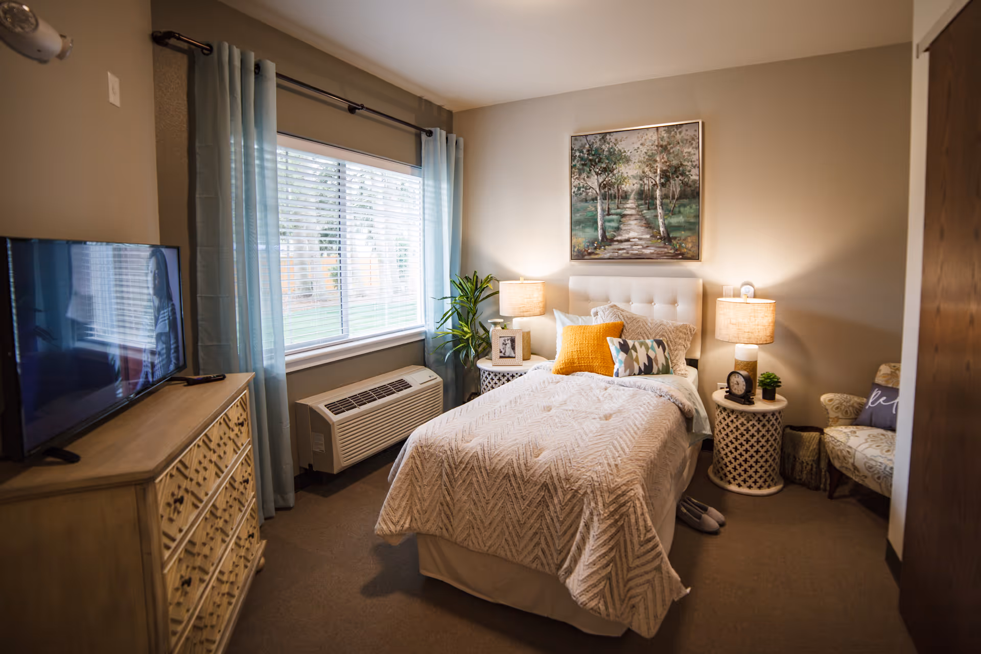A cozy bedroom in a senior living facility featuring a single bed with white and patterned bedding, multiple pillows including a yellow one, two bedside tables with lamps, a framed picture, a clock, and a small plant. There is a large window with blue curtains, a dresser with a TV on top, a patterned armchair with a pillow, and a painting of a tree-lined path hanging above the bed.