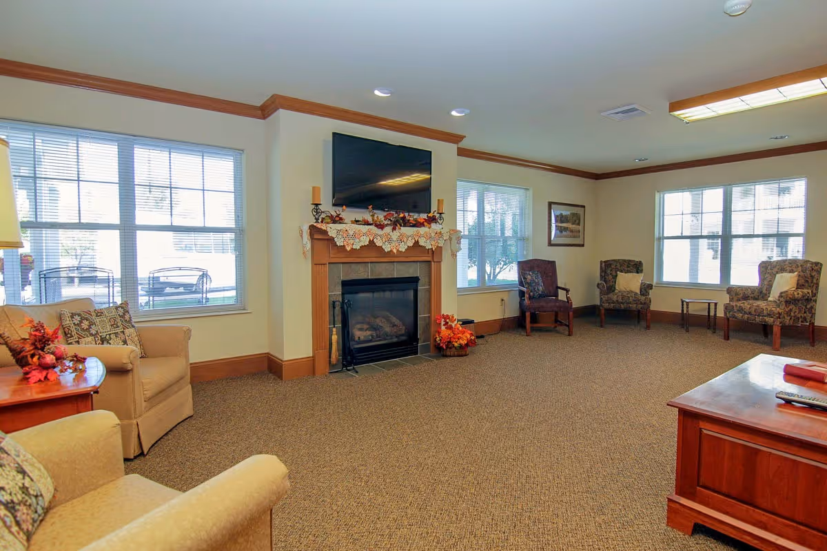 A cozy senior living room with beige walls and carpet, featuring a fireplace with a TV mounted above it. The fireplace mantel is decorated with a lace runner and autumn-themed decor. There are several armchairs arranged around the room, with large windows letting in natural light. A wooden coffee table is visible in the foreground.
