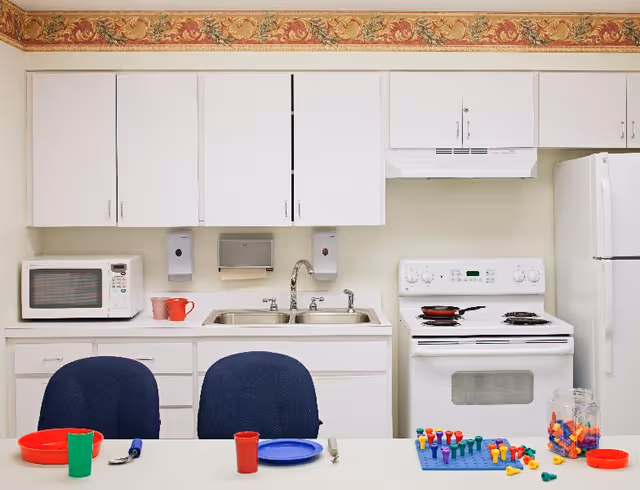 Institutional-style kitchen with white cabinets, sink, stove and refrigerator, and a table set with colorful plates, cups and toys.