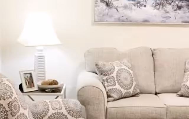 A cozy living room area with a beige couch and matching armchair featuring patterned cushions. A white side table holds a lamp, a framed photo, and a bowl with decorative items. A framed artwork hangs on the wall above the couch.