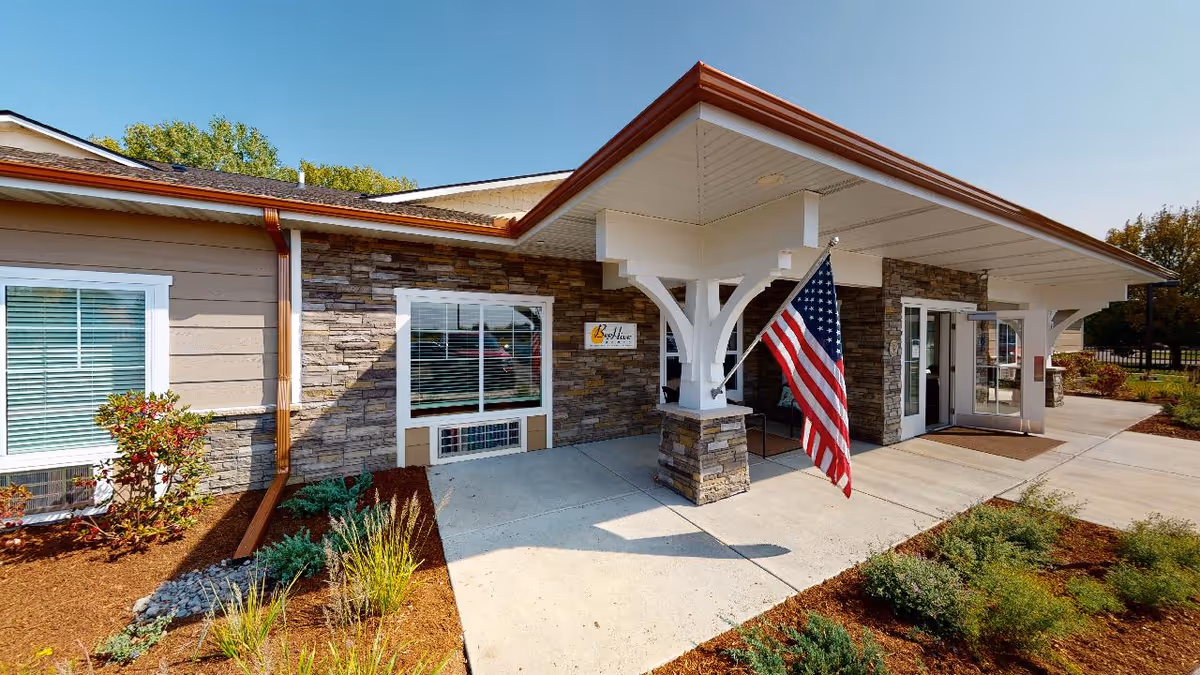 Exterior view of Beehive Homes Of Lakeville building entrance with stone and beige siding, a covered porch supported by white beams, an American flag mounted on a post, and landscaped plants along the walkway.