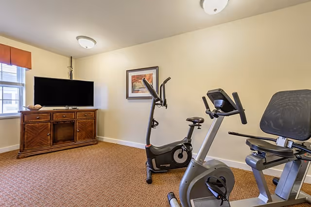 A small exercise room with two stationary bikes facing a wooden cabinet with a flat-screen TV on top. The room has beige walls, a window with a red valance, a framed picture on the wall, and carpeted flooring.