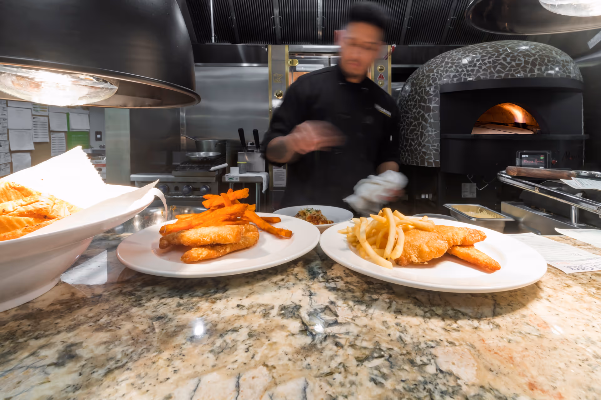 A kitchen scene with a chef preparing food behind a counter. Two plates of fried fish with fries are placed on the counter, one with sweet potato fries and the other with regular fries. A pizza oven is visible in the background.