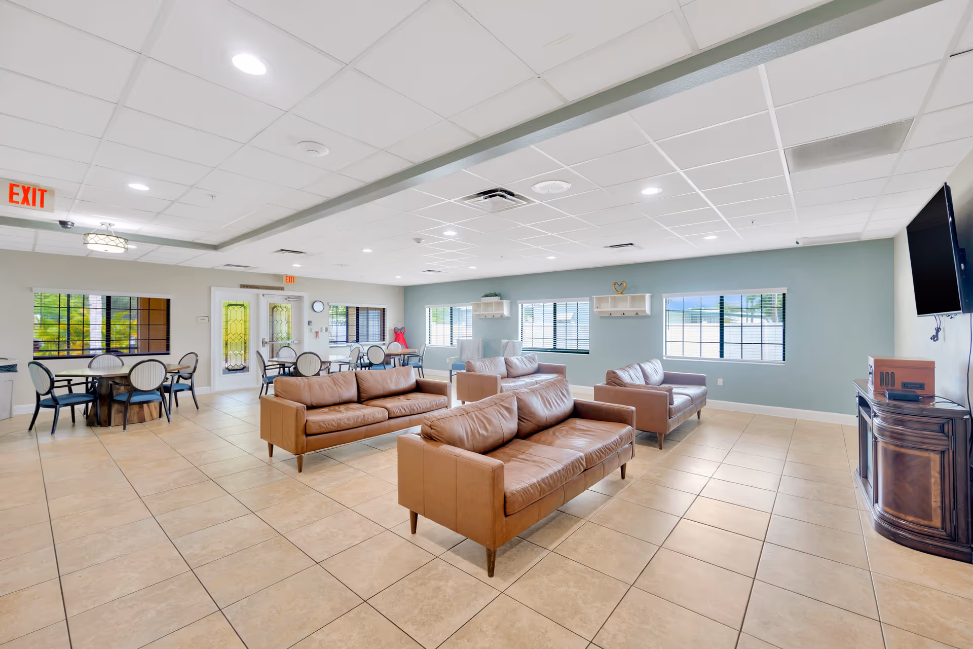 Spacious senior living common room with leather sofas, dining tables, large windows and a wall-mounted TV.