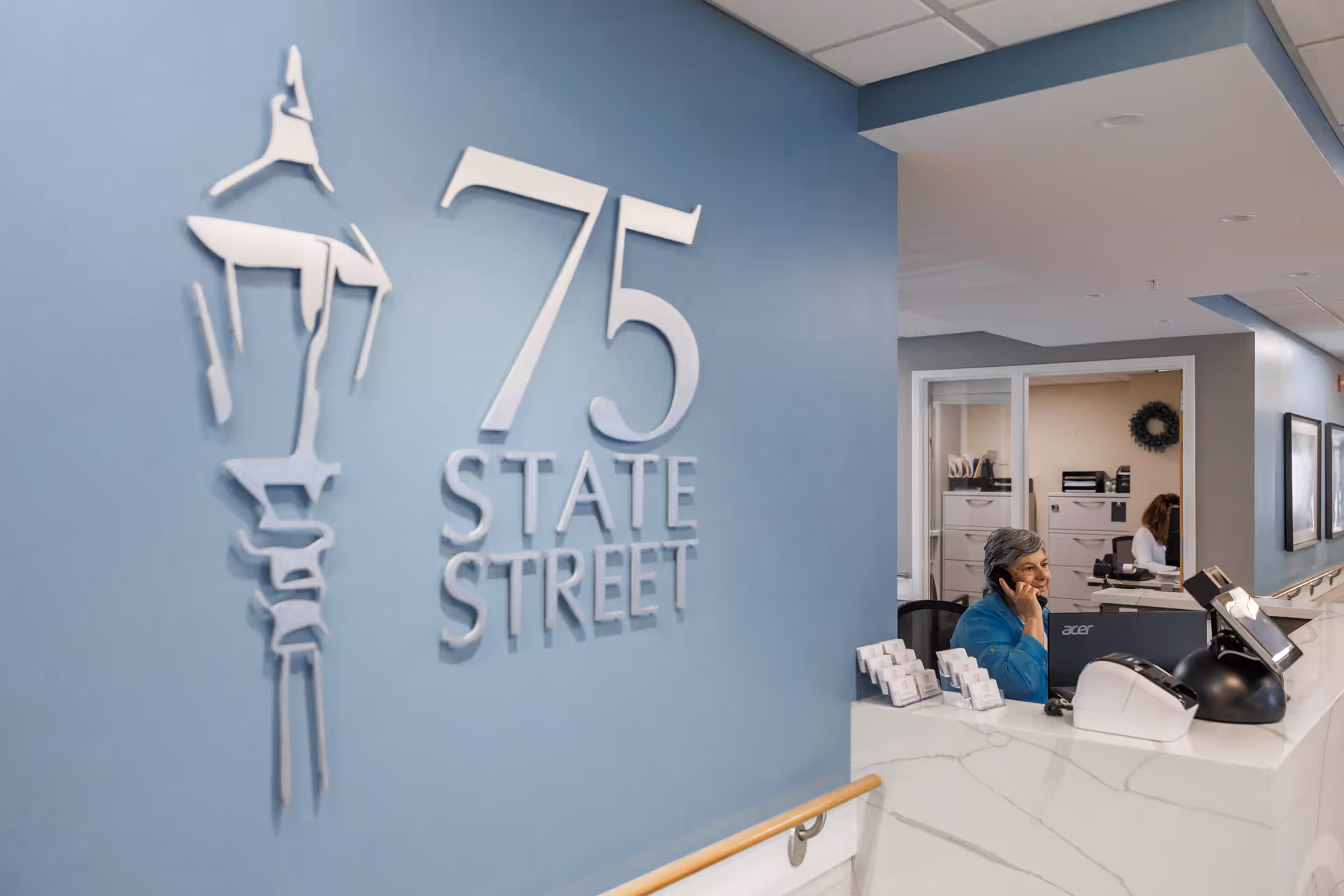 Reception area of 75 State Street facility with a woman sitting behind a white marble counter, talking on the phone. The wall behind her is blue with a silver 75 State Street logo. The area looks clean and professional with office equipment and another person working in the background.