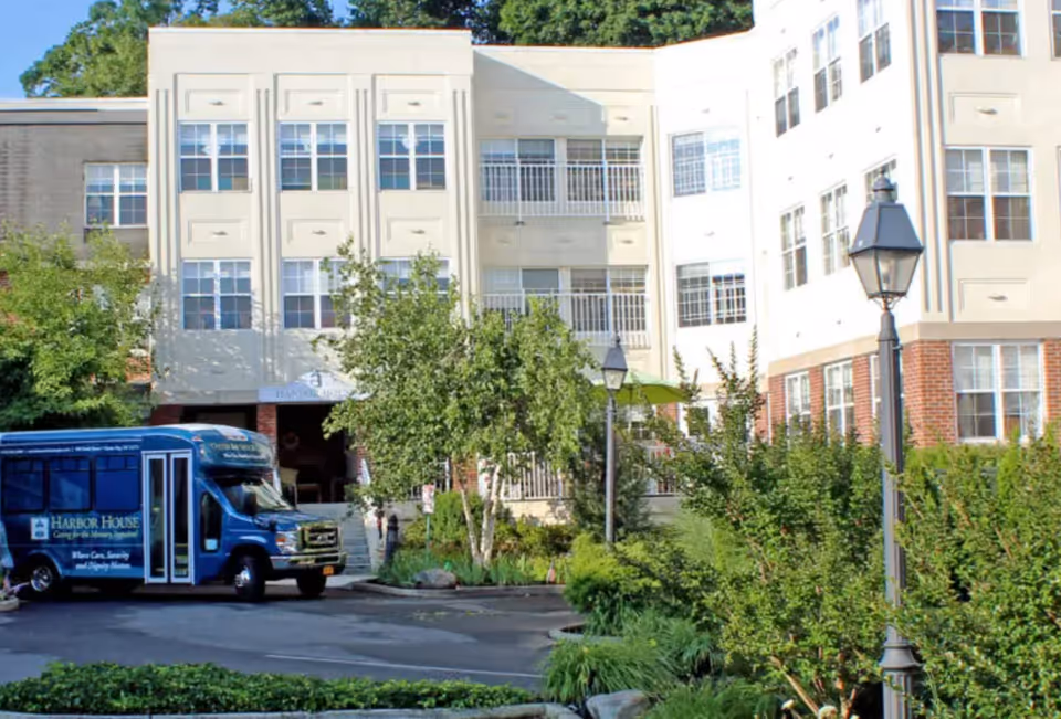 Exterior view of Harbor House Assisted Living facility showing a multi-story building with numerous windows, surrounded by greenery and trees. A blue Harbor House shuttle bus is parked in front of the entrance, and there are street lamps and landscaped bushes along the driveway.