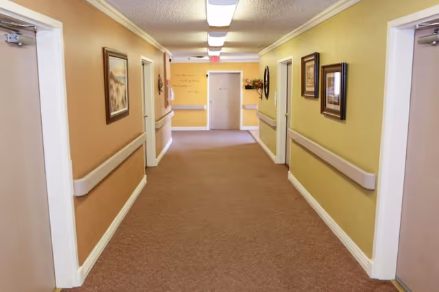 A well-lit hallway in a senior living facility with beige and yellow walls, carpeted floor, handrails on both sides, several closed doors, framed pictures on the walls, and an exit door at the end.